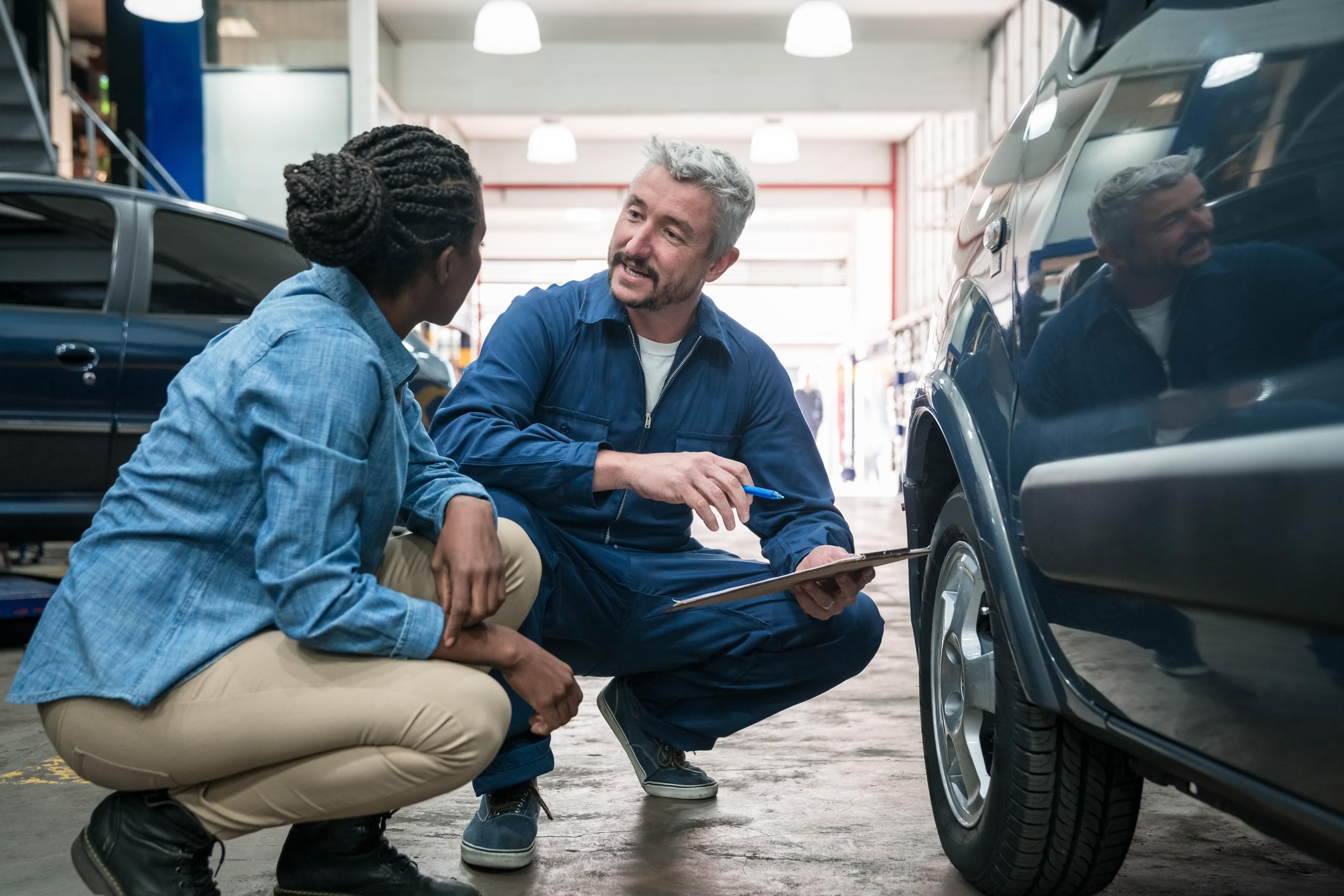 Two people are crouched down on the floor, one is a mechanic showing the other something on a car